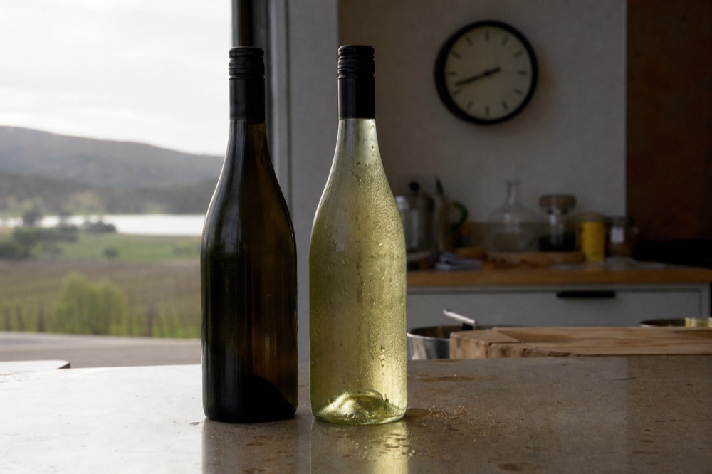Two wine bottles on a kitchen bench with condensation beading on the chilled one and a clock visible behind