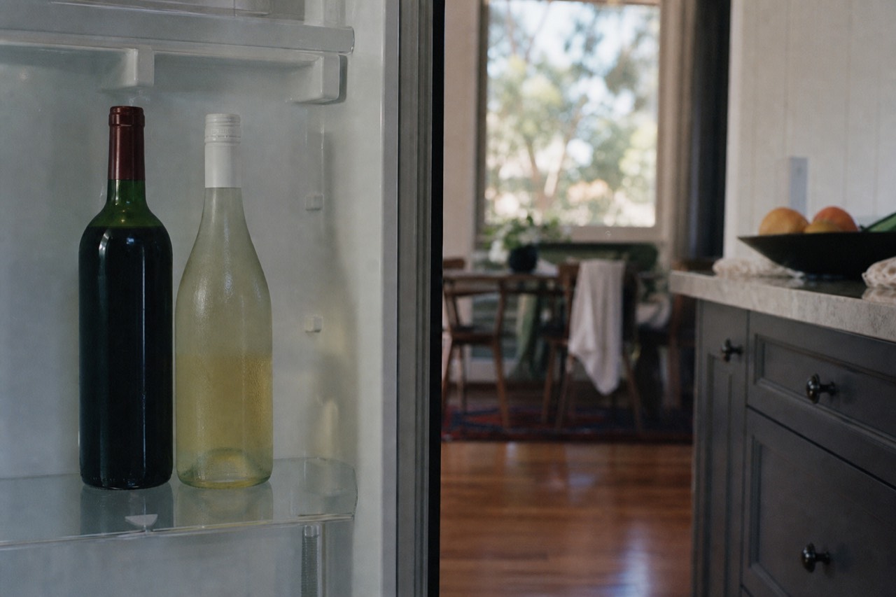 Bottle of red wine standing on a refrigerator shelf beside a chilled white bottle on a kitchen bench