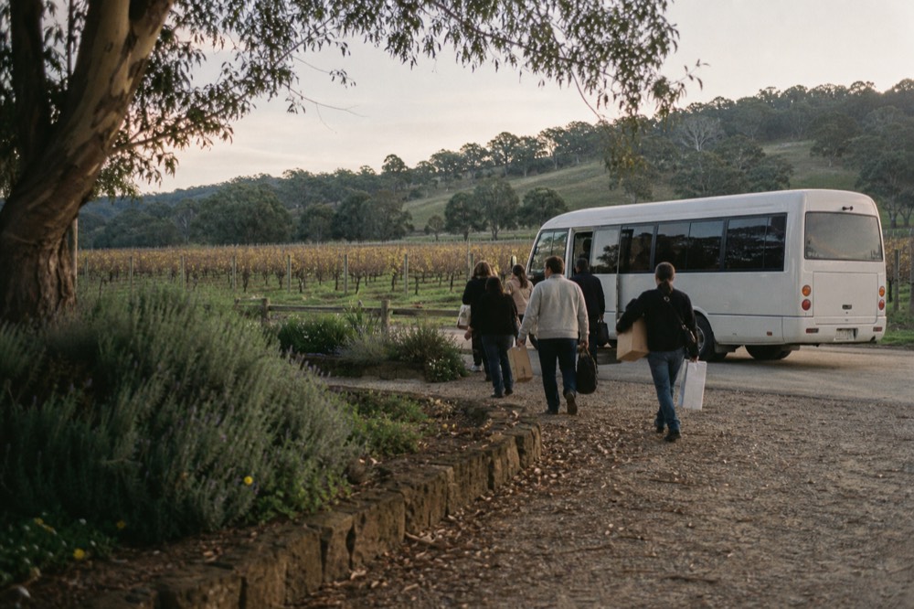 A tour minibus parked at the edge of a vineyard in late afternoon light, passengers walking back toward it carrying wine boxes