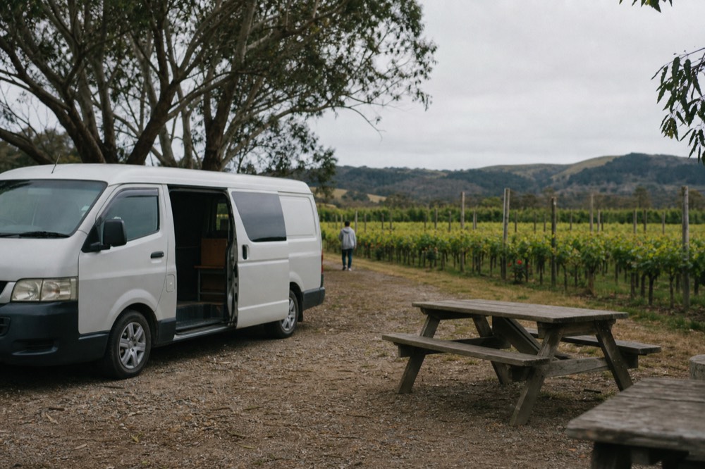 Small van parked on a gravel driveway beside a vineyard with its side door open and a picnic bench nearby