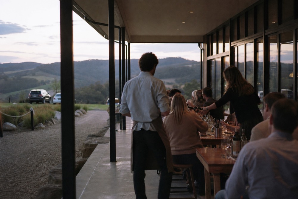 An open cellar door tasting room with two staff pouring for visitors seated at a long communal table beside tall windows