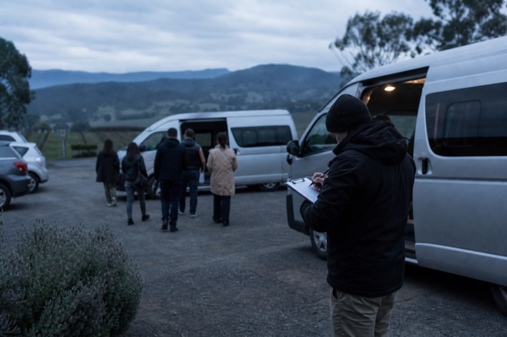 A host writing on a clipboard beside an open van door in a winery car park, guests boarding in the background, overcast morning