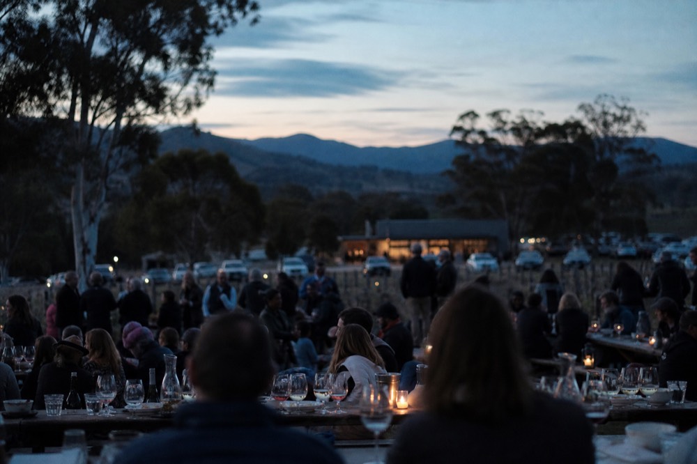 Outdoor city festival setting with long communal tables, wine glasses catching the light and a crowd blurred in the background, no faces in focus