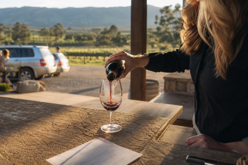 Cellar-door staff member pouring a small measure of red wine at a stone counter with a printed tasting list beside the glass
