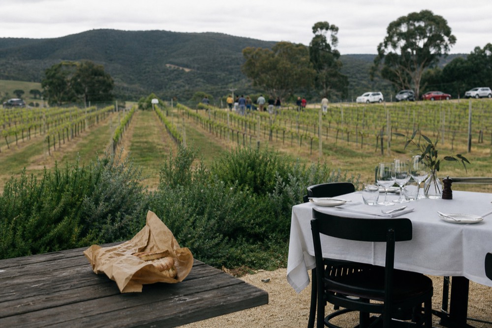 Two contrasting tables side by side, a paper-wrapped lunch on a bench and a set restaurant table with linen overlooking vines