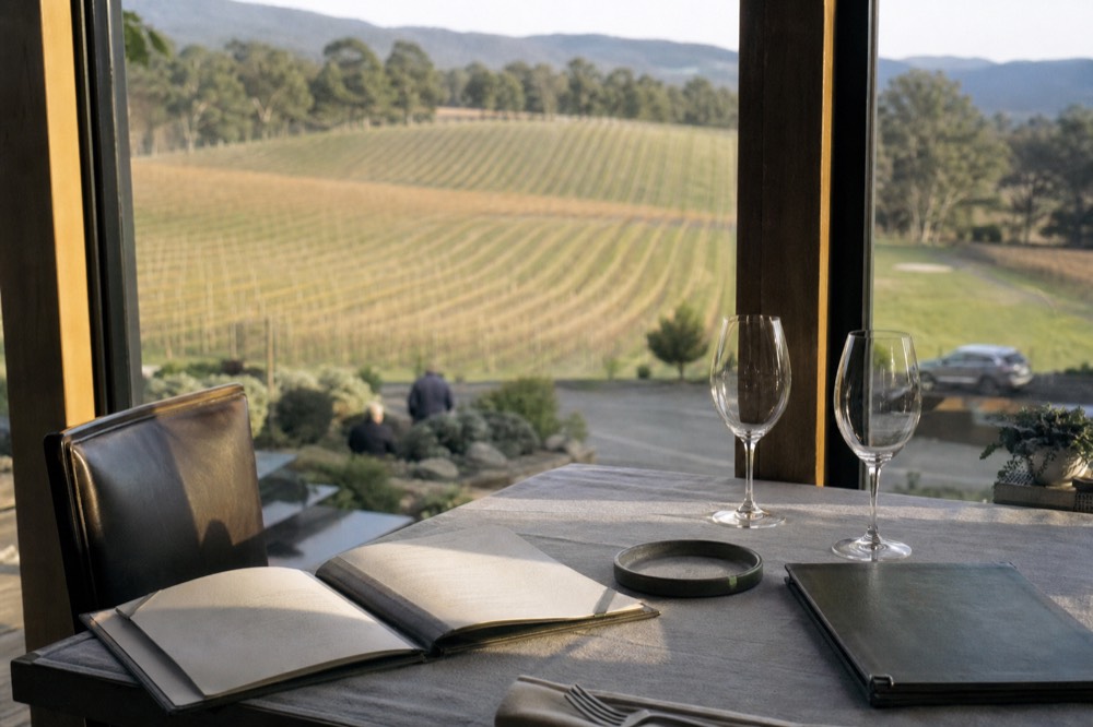 Open restaurant menu on a linen-set table beside a wine list and two empty glasses in a vineyard dining room